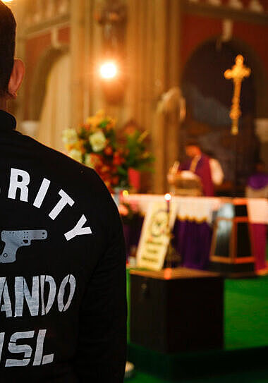 Ein Mensch von hinten fotografiert: Auf seinem Shirt steht „SECURITY”. Im Hintergrund ist der Altar einer Kirche zu sehen.