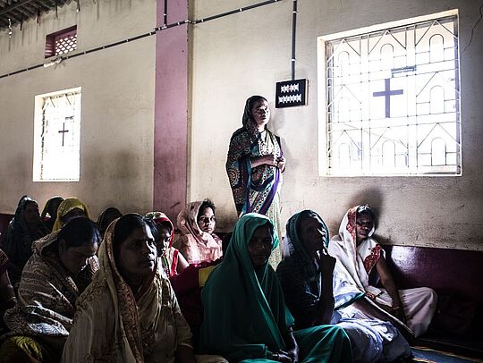 Christ/-innen in einer Kirche in Indien. Sie sitzen auf dem Boden. Der Raum wird durch Licht aus drei Fenstern erhellt.