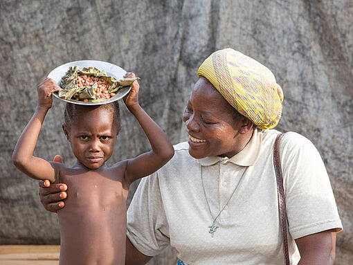 Schwester Angélique sitzt neben einem Kind, das einen Teller auf dem Kopf trägt.