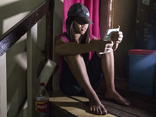 Ein philippinisches Mädchen sitzt auf einer Holztreppe mit einem Handy in der Hand. 