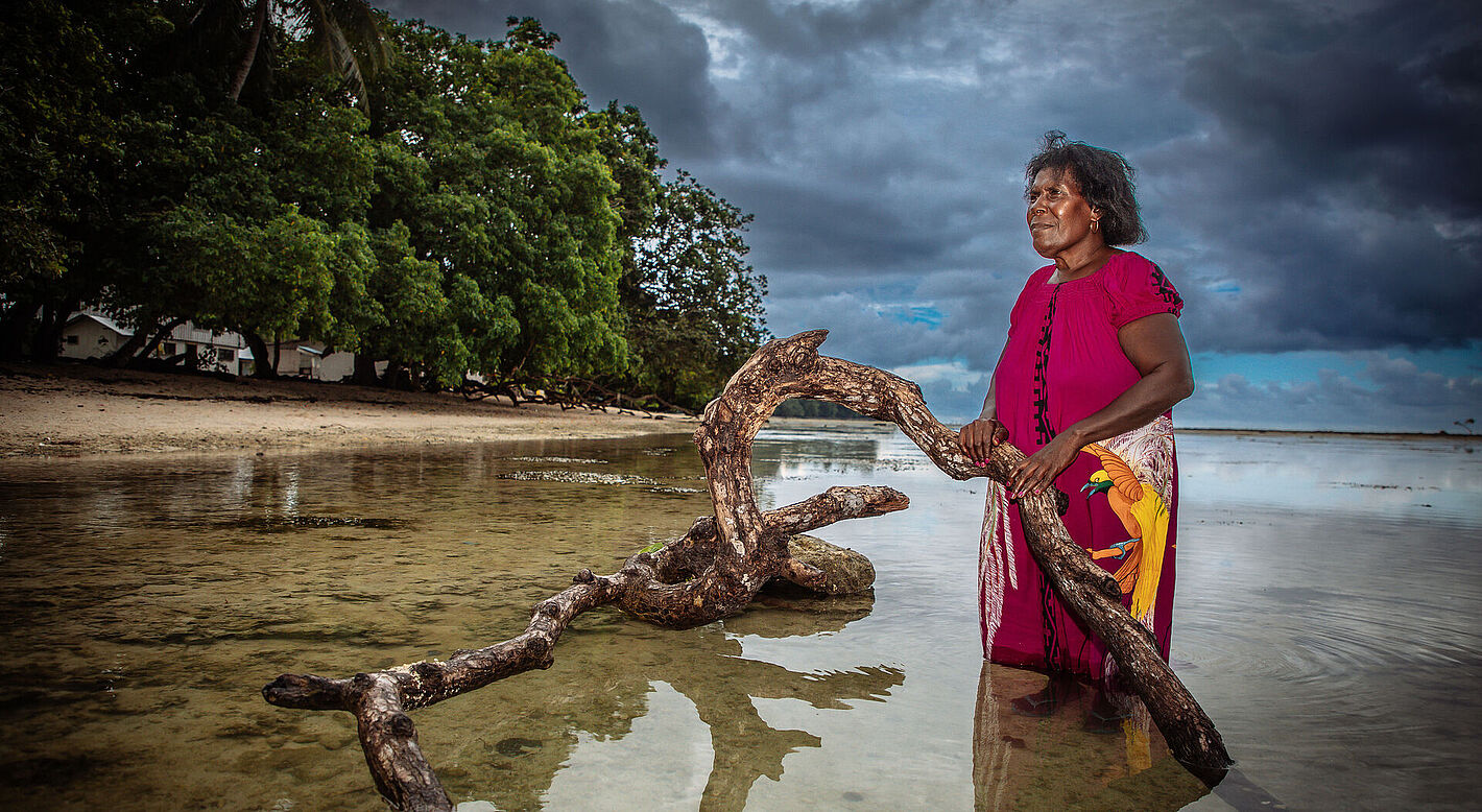 Helen Samu Hakena ist eine Organisatorin und Aktivistin für Frieden und Frauenrechte aus Bougainville, Papua-Neuguinea. 1992 war sie Mitbegründerin der Leitana Nehan Women's Development Agency, um zur Wiederherstellung des Friedens auf der Insel beizutragen. Sie ist auch die Präsidentin der Katholischen Frauengemeinschaft der Erzdiözese Bougainville. Das Foto zeigt sie am Strand vor ihrem Haus. Die toten Bäume zeugen davon, dass hier einmal festes Land war. Sie steht auf dem Grundstück des Hauses ihres Sohnes, das schon vom Meer verschlungen wurde. Die Bewohner fühlen sich durch den Anstieg des Meeresspiegels und den Klimawandel massiv bedroht.