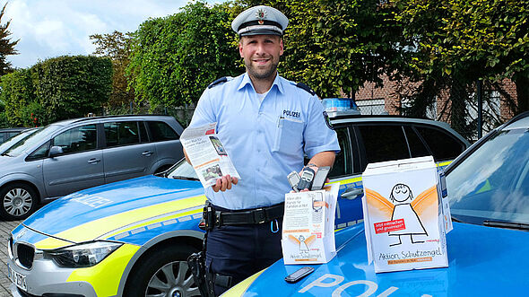 Ein Polizist in Münster spendet sein altes Handy und steht mit Infomaterialien an einem Polizeiwagen.