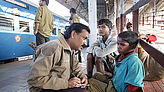 An einem Bahnhof in Indien spricht ein Mann mit einem kleinen Jungen und macht sich Notizen.
