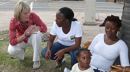 Gundula Gause sitzt in der Hocke und spricht mit der Südafrikanerin Zanele und einer Frau mit Kind.